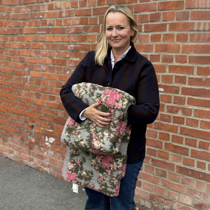 En dame står foran murvegg og holder frem et pledd